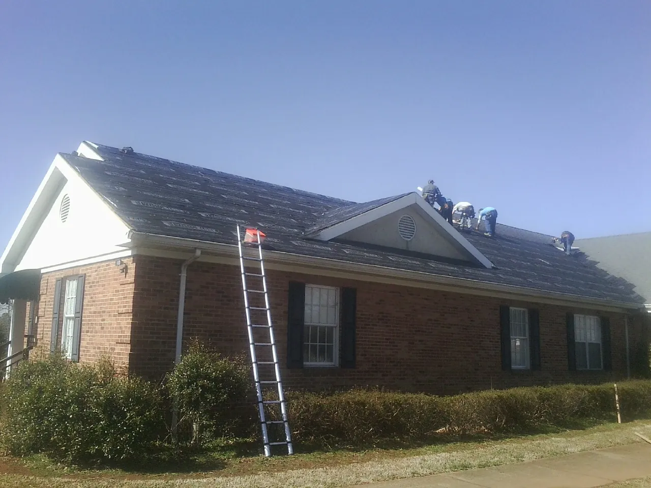 Crew tearing shingles off a brick ranch roof with a ladder set — Emergency Roof Repair work by our crew in Jamaica Plain