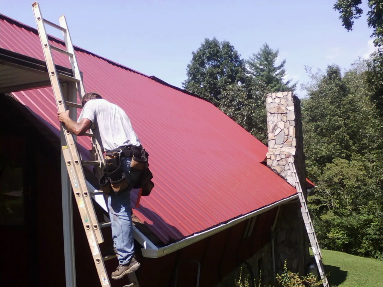 Roofer on a ladder beside a red metal roof and stone chimney — Soffit & Fascia Repair by Roofing Brothers in Jamaica Plain